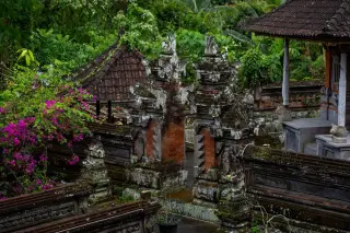 Ilustrasi: Pintu gerbang pura berarsitektur khas Bali dikelilingi pepohonan hijau dan bunga merah muda.