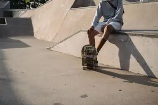 Ilustrasi: Seseorang duduk di tepi rintangan skatepark beton dengan papan skateboard di depannya.