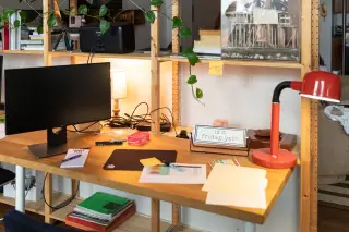 Ilustrasi: Meja kerja kayu dengan monitor, lampu meja merah, dan beberapa dokumen di atasnya.