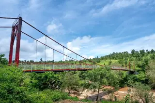 Ilustrasi: Jembatan gantung berstruktur merah membentang di atas sungai beraliran deras di kawasan perbukitan hijau.