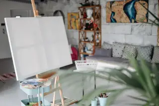 Ilustrasi: Kanvas putih di atas kuda-kuda dan palet cat di dalam studio seni yang tertata rapi.