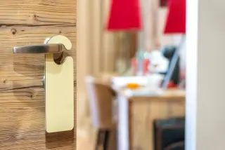 Ilustrasi: Gagang pintu kamar hotel berbahan kayu dengan gantungan tanda kosong dan latar interior kamar.