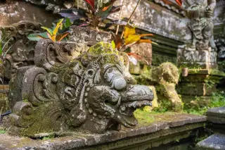 Ilustrasi: Ukiran batu tradisional Bali berlumut di area pura atau pekarangan dengan tanaman hijau.