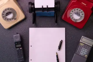 Ilustrasi: Tata letak telepon putar antik, pena, dan kertas catatan bergaris di atas meja.