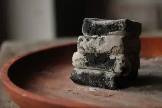 Ilustrasi: Tumpukan briket arang berbalur abu di atas piring kayu cokelat.