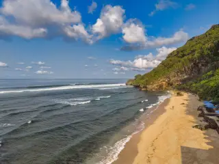Ilustrasi: Pemandangan pantai berpasir dan tebing hijau di bawah langit biru cerah berawan.