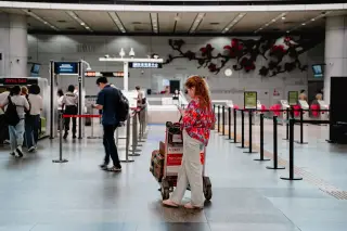 Ilustrasi: Seorang wanita berdiri memegang ponsel di dekat troli bagasi di area terminal bandara.