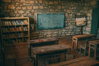 Ilustrasi: Ruang kelas berdinding batu dengan meja kayu kosong, papan tulis, dan rak buku.