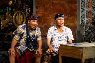 Ilustrasi: Dua pria mengenakan udeng dan kain adat duduk bersantai di dekat bangunan batu bata.