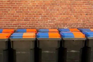 Ilustrasi: Deretan tempat sampah beroda dengan penutup berwarna oranye dan biru di depan dinding bata merah.