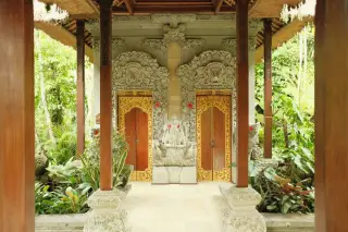 Ilustrasi: Bangunan berukir khas Bali dengan pintu kayu berhias emas di antara tanaman tropis.