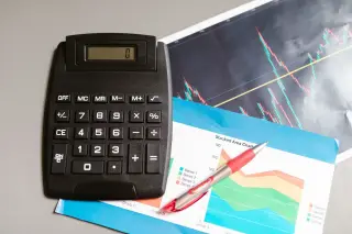 Ilustrasi: Kalkulator hitam, pulpen, dan kertas grafik keuangan di atas meja.