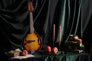 Ilustrasi: Sebuah mandolin bersandar di meja bersama apel, lilin, dan buku berlatar tirai hitam.