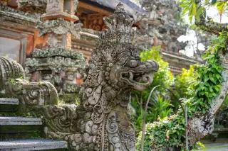 Ilustrasi: Patung naga batu berukir khas Bali di tangga sebuah pura dengan latar pepohonan.