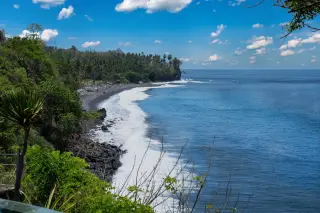 Ilustrasi: Garis pantai berbatu dan bukit hijau dengan deburan ombak berbusa putih di bawah langit cerah.