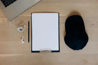 Ilustrasi: Papan klip dengan kertas kosong, pensil, laptop, earphone, dan topi hitam di atas meja kayu.