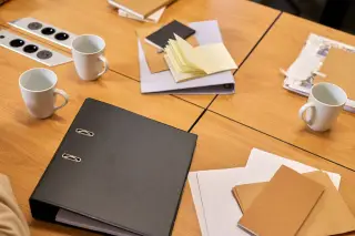 Ilustrasi: Buku binder hitam, cangkir kopi, dan tumpukan dokumen di atas meja rapat kayu.
