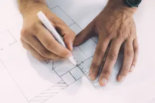 Ilustrasi: Sepasang tangan menggambar denah arsitektur menggunakan pena dan penggaris.