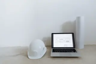 Ilustrasi: Helm proyek putih, laptop dengan gambar denah, dan gulungan kertas di lantai.