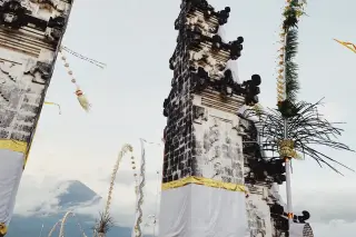 Ilustrasi: Gapura candi bentar khas Bali berhias kain dan penjor dengan latar gunung berawan.