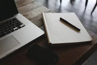 Ilustrasi: Buku catatan bergaris kotak, pulpen, ponsel, dan laptop di atas meja kayu.