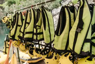Ilustrasi: Deretan rompi pelampung hijau muda dan helm kuning untuk arung jeram tergantung di luar ruangan.