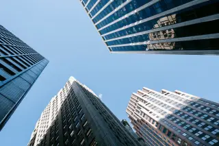Ilustrasi: Sudut pandang rendah deretan gedung pencakar langit dengan fasad kaca dan beton berlatar langit biru.