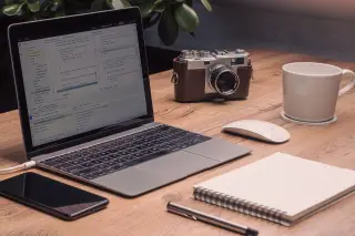 Ilustrasi: Laptop, kamera antik, buku catatan, dan cangkir kopi di atas meja kayu.