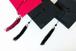 Ilustrasi: Tiga topi toga wisuda berwarna merah dan hitam dengan rumbai di atas latar putih.