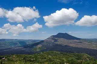 Ilustrasi: Pemandangan Gunung Batur dengan hamparan lava hitam di bawah langit biru berawan.