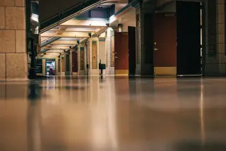 Ilustrasi: Lorong gedung bertiang batu yang sepi dengan lantai mengilap dan deretan pintu kayu terbuka.