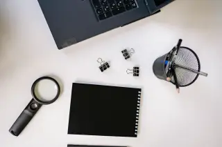 Ilustrasi: Tampilan atas meja kerja dengan laptop, buku catatan hitam, penjepit kertas, dan kaca pembesar.