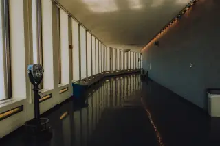 Ilustrasi: Lorong panjang yang sepi dengan deretan jendela besar dan teropong koin di dekatnya.