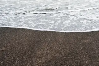 Ilustrasi: Buih ombak putih menyapu permukaan pasir pantai berwarna hitam.