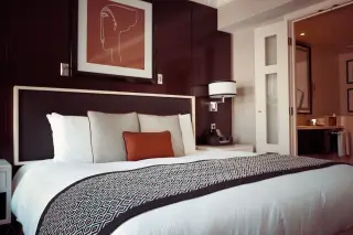 Ilustrasi: Kamar tidur hotel dengan tempat tidur rapi, hiasan dinding, dan pintu kamar mandi terbuka.