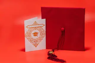 Ilustrasi: Topi wisuda merah dan kartu ucapan wisuda berlatar belakang merah cerah.