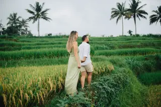 Ilustrasi: Sepasang turis berjalan bergandengan tangan di pematang sawah berundak dengan pohon kelapa.