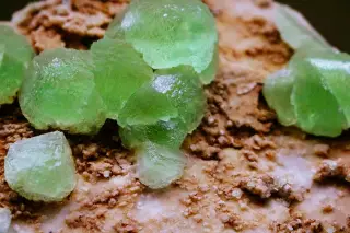 Ilustrasi: Bongkahan kristal mineral berwarna hijau cerah di atas permukaan batuan berpasir.