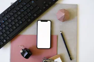 Ilustrasi: Tampilan flat lay meja kerja dengan ponsel berlayar putih polos, papan ketik, dan alat tulis.