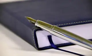 Ilustrasi: Pena bulu logam berwarna perak diletakkan di atas buku agenda bersampul biru tua.