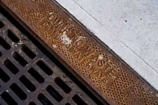 Ilustrasi: Kisi besi drainase berkarat dengan tulisan peringatan di samping trotoar beton.