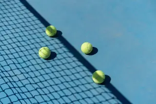 Ilustrasi: Empat bola tenis berada di lapangan biru dekat bayangan jaring.