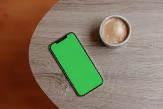 Ilustrasi: Ponsel pintar berlayar hijau dan secangkir kopi di atas meja bundar bermotif kayu.