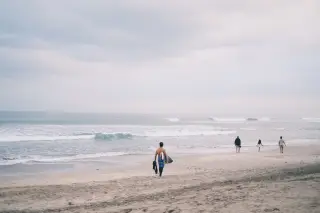Ilustrasi: Seseorang membawa papan selancar berjalan di tepi pantai berpasir menuju laut berombak di bawah langit mendung.