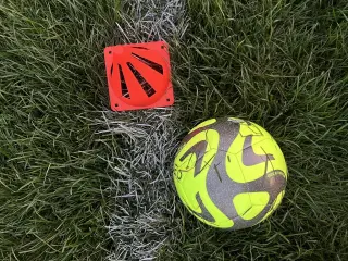 Ilustrasi: Sebuah bola sepak hijau neon dan kerucut latihan oranye di atas rumput lapangan bergaris putih.