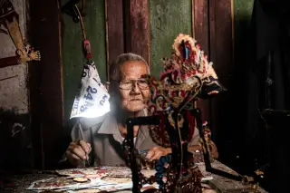Ilustrasi: Seorang pria lansia berkacamata sedang mewarnai wayang kulit di bawah lampu kecil.
