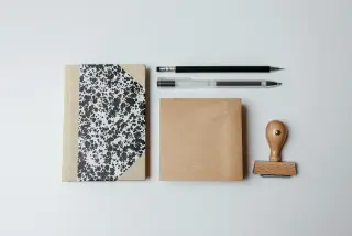 Ilustrasi: Buku catatan, alat tulis, kertas cokelat, dan stempel kayu tertata di atas meja putih.