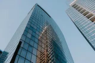 Ilustrasi: Gedung pencakar langit modern berdinding kaca dengan pantulan gedung lain di bawah langit cerah.
