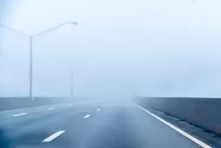 Ilustrasi: Jalan raya beraspal yang kosong dan diselimuti kabut tebal dengan tiang lampu jalan.