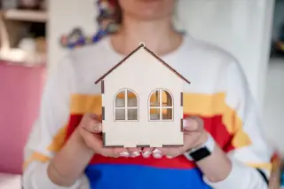 Ilustrasi: Seseorang memegang miniatur rumah kayu sederhana dengan kedua tangan.